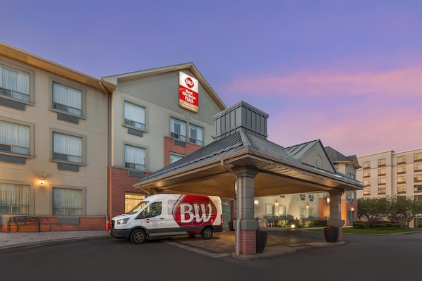Un hotel con acceso cubierto, una camioneta con la marca 'BW' y un letrero que indica los servicios de 'Best Western Plus'.
