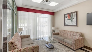 A modern hotel room with a glass coffee table, two patterned sofas, a large window with sheer curtains, and a framed abstract painting on the wall.