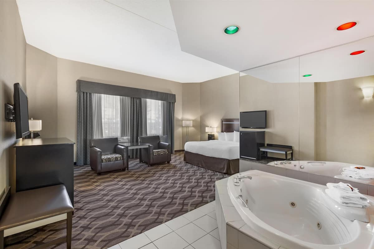 Habitación de hotel con una cama grande, zona de estar con una silla y una mesita, televisor, baño con bañera y espejo.