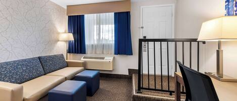 A hotel room with a sofa, two ottomans, a desk with a chair, a lamp, a window with curtains, an air conditioning unit, and a door.