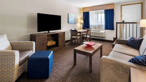 Ett hotellrum med en platt-TV, en soffa, ett soffbord, ett matbord med stolar och ett fönster med gardiner.