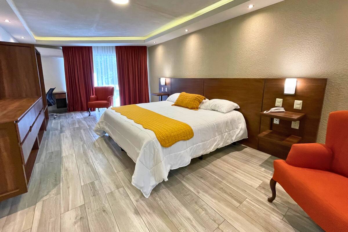 Una habitación de hotel con una cama grande, un armario de madera, un escritorio con una silla, un sillón rojo y una mesita de noche con un teléfono.