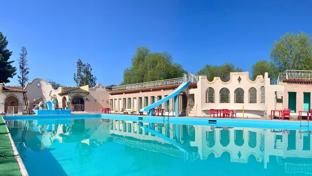 En swimmingpool med en vattenrutschkana, röda stolar och en byggnad med välvda fönster och en balkong.