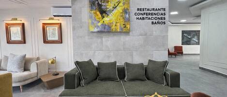 A modern lounge area with a sofa, framed artwork, and a wall with text indicating amenities like a restaurant, conference rooms, accommodations, and a pool.