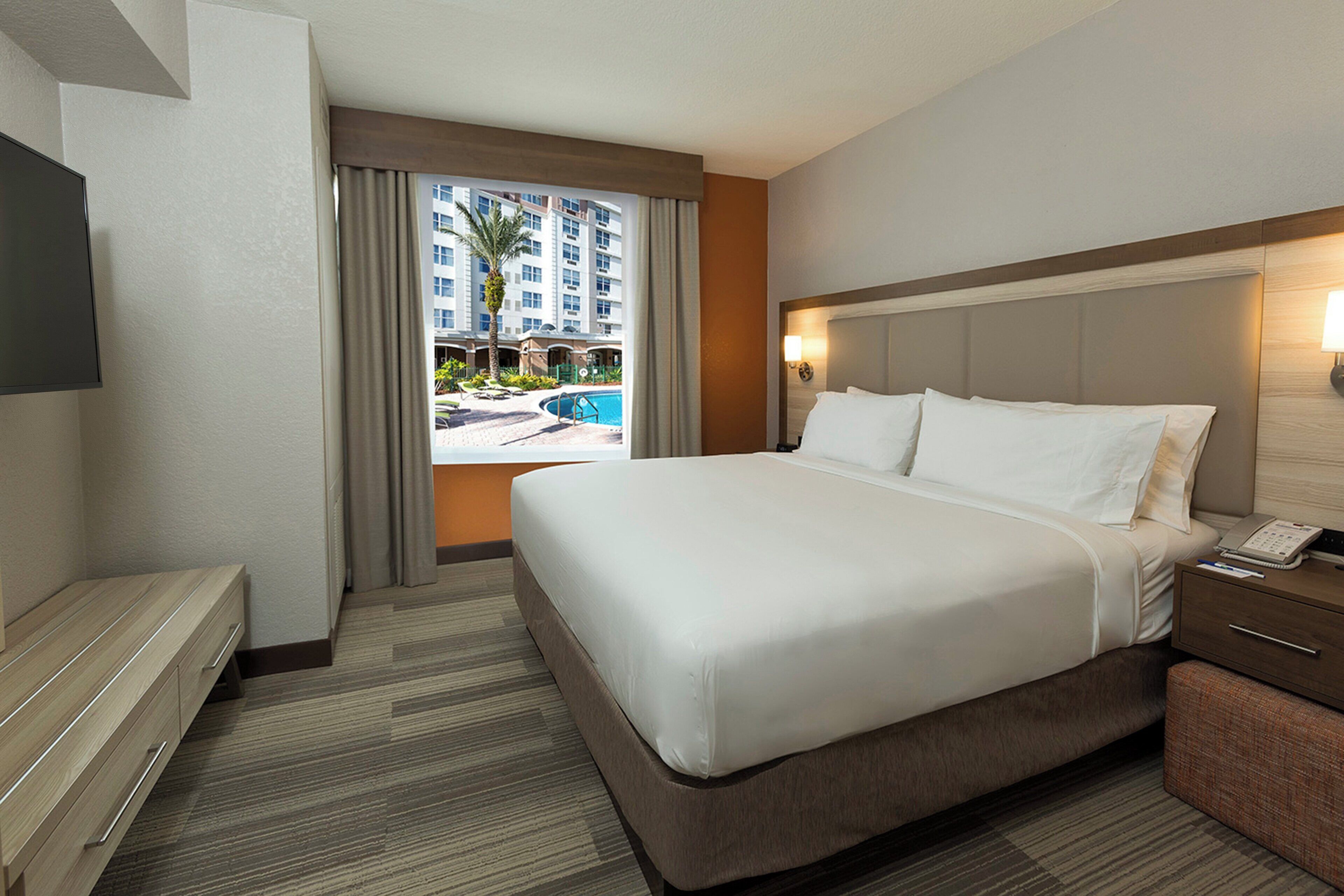 A hotel room with a large bed, a desk, a chair, a TV, and a view of a swimming pool and buildings.