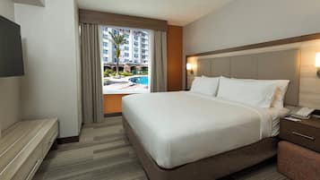 A hotel room with a large bed, a desk, a chair, a TV, and a view of a swimming pool and buildings.