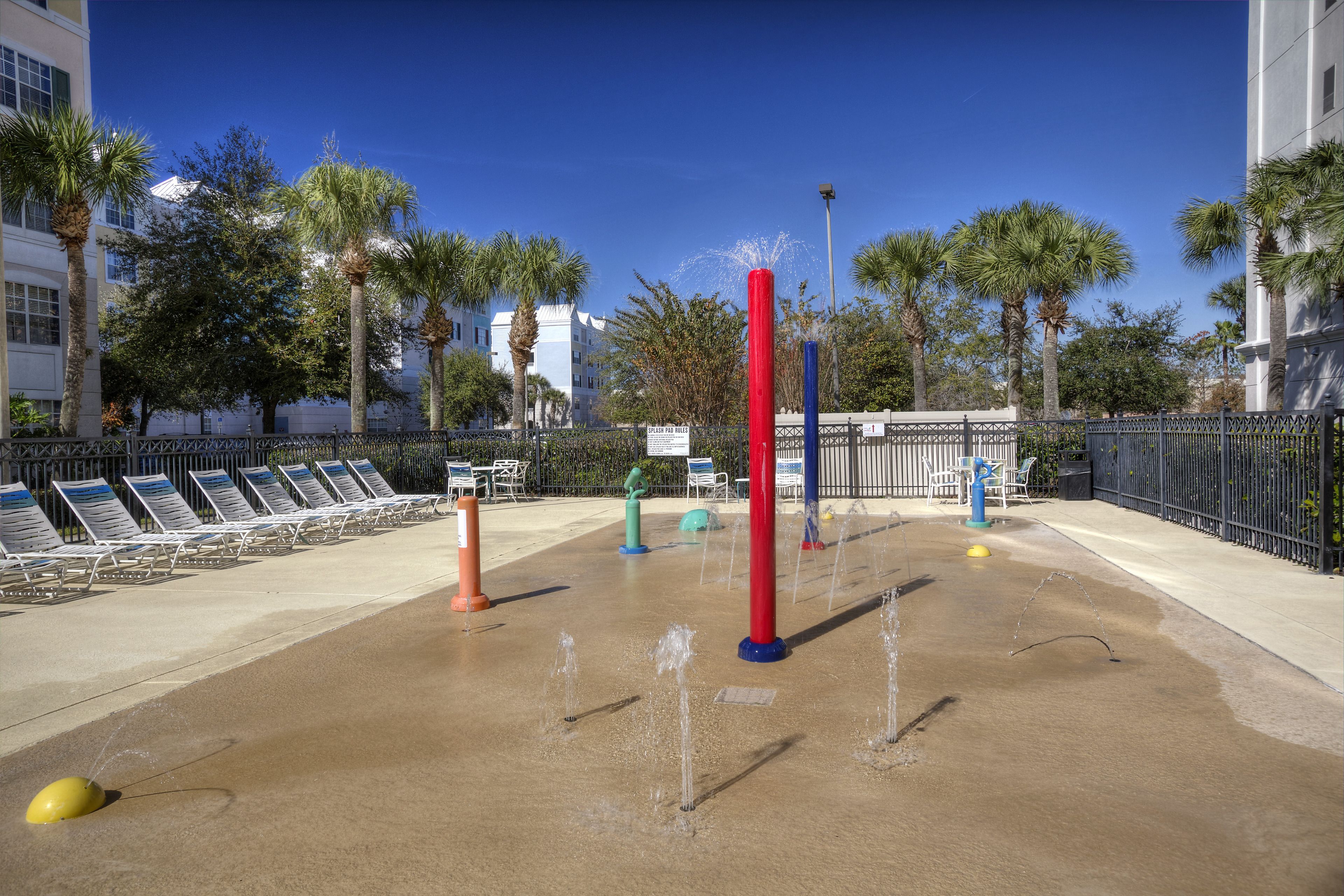 Un espace de jeux aquatiques doté de plusieurs jets d’eau, entouré de chaises longues et de palmiers.