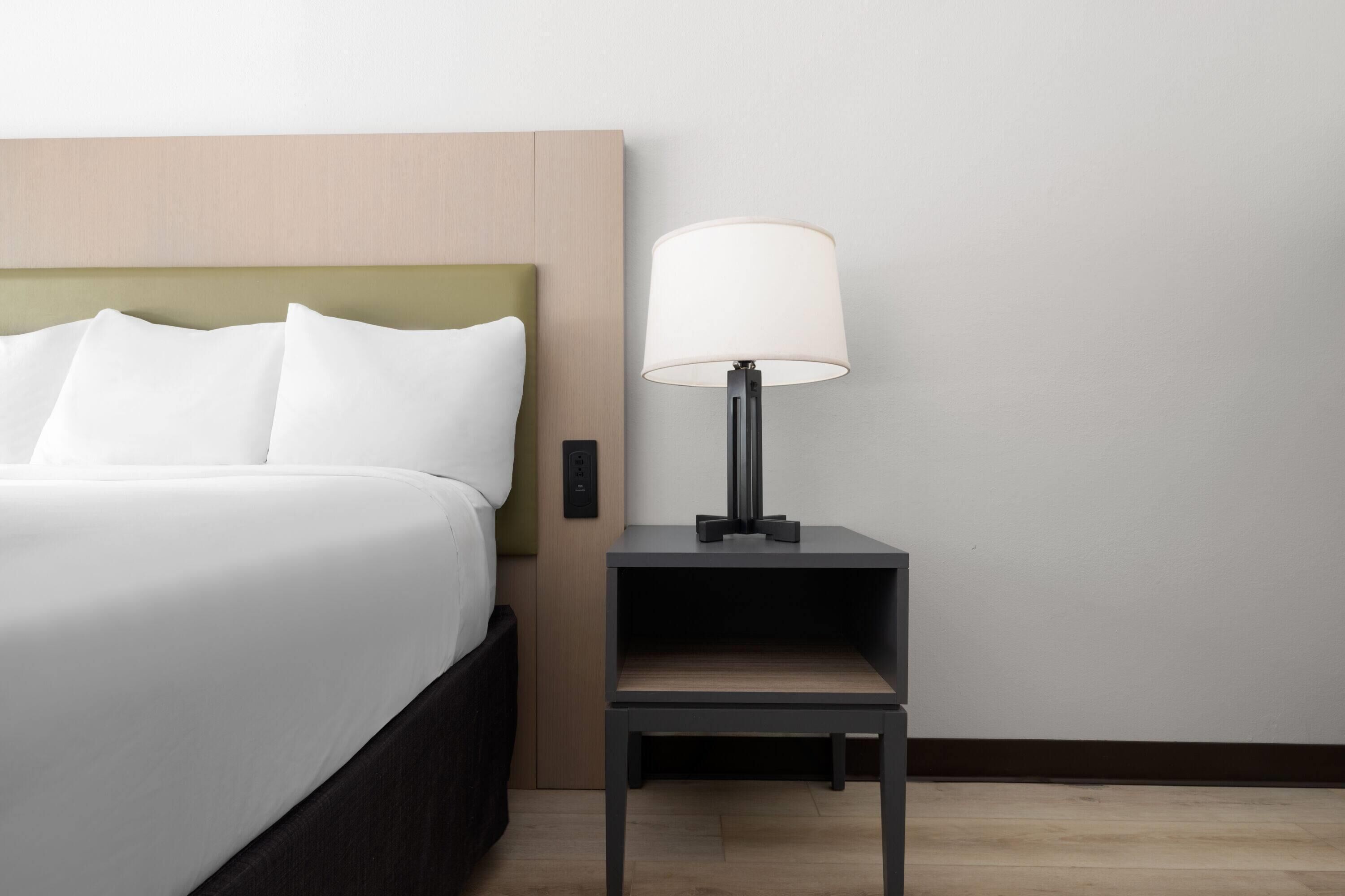 A neatly made bed with white linens and a green headboard, a small bedside table with a lamp, and a plain wall.
