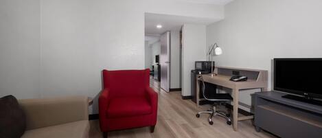A modern hotel room with a beige sofa, a red armchair, a wooden desk with a computer, a TV, and a flat-screen TV on a stand.