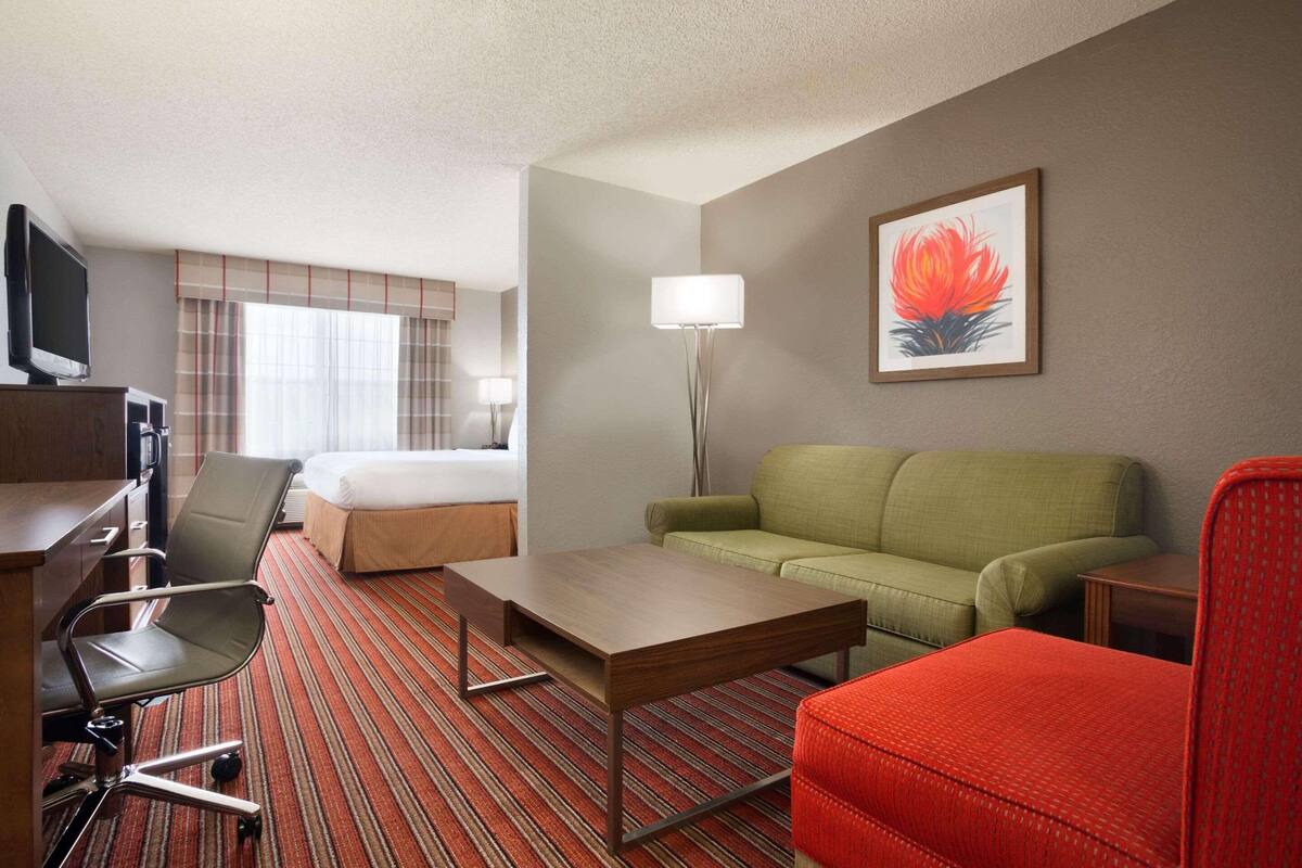 A hotel room with a bed, a green sofa, a red armchair, a wooden coffee table, a desk with a chair, a TV, and a painting on the wall.