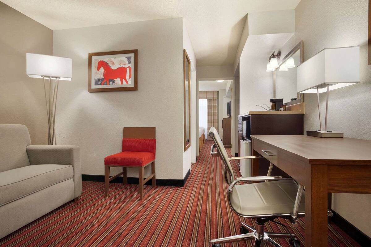 A hotel room with a sofa, a red chair, a desk with a computer, a lamp, and a framed picture on the wall.
