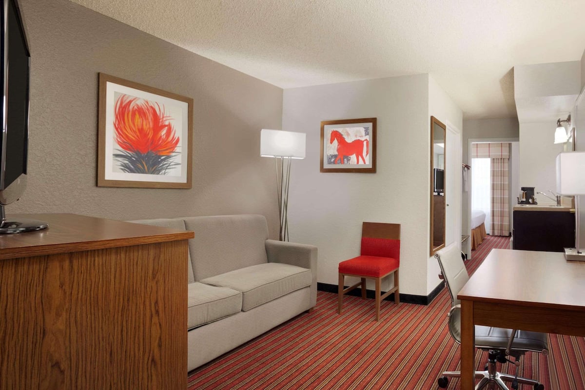 A hotel room with a wooden desk, a beige sofa, a red chair, a dining table, and two framed artworks on the walls.