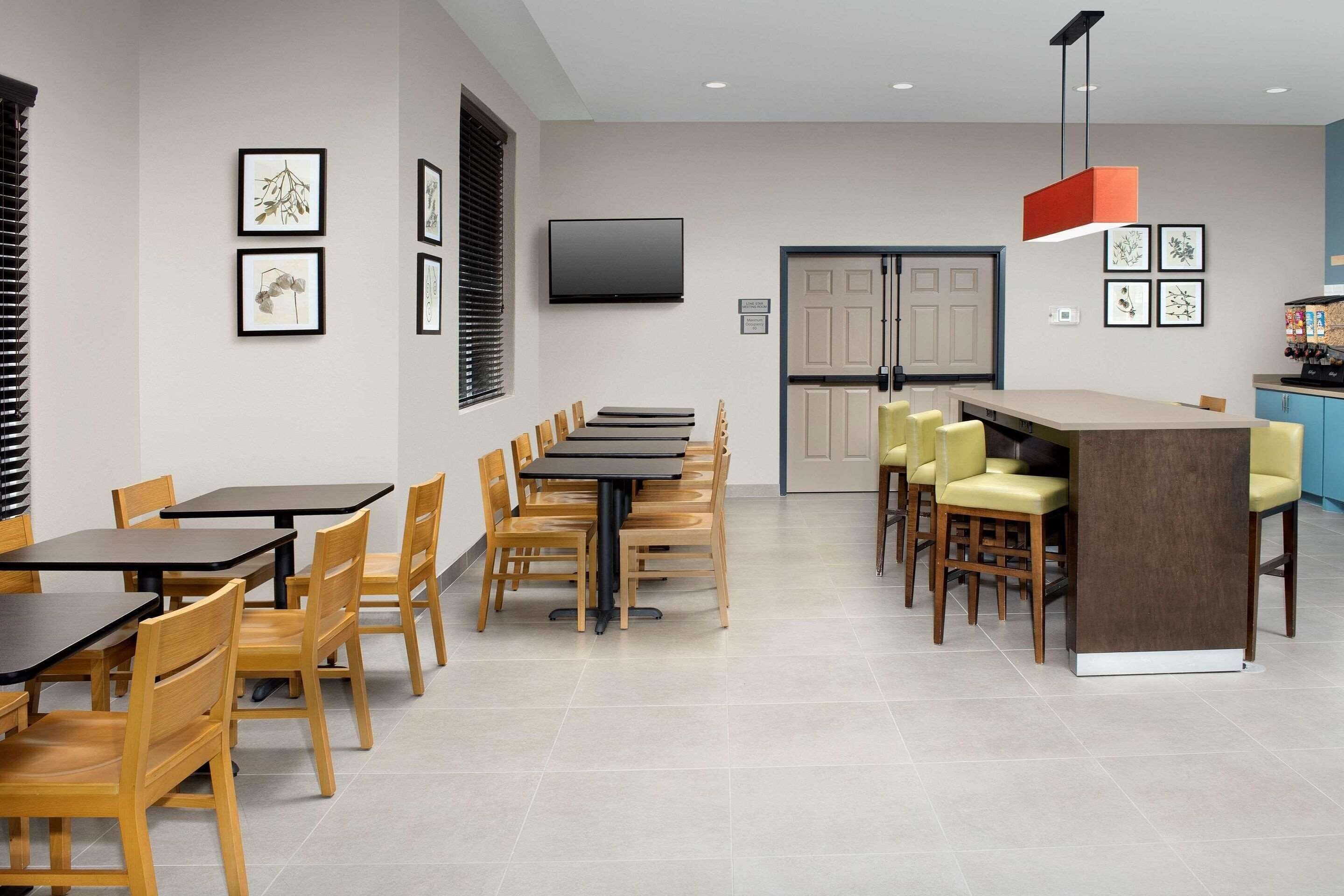 A modern dining area with wooden tables and chairs, a flat-screen TV mounted on the wall, and a kitchen area with a bar.
