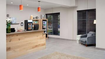 Una moderna zona de recepción con un mostrador de madera, un sillón gris y un amplio ventanal con persianas.