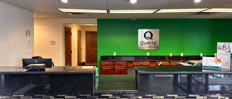 Zona de recepción de un hotel con una pared verde, un letrero que dice "Quality by Choice Hotels" y un mostrador de recepción con una computadora.