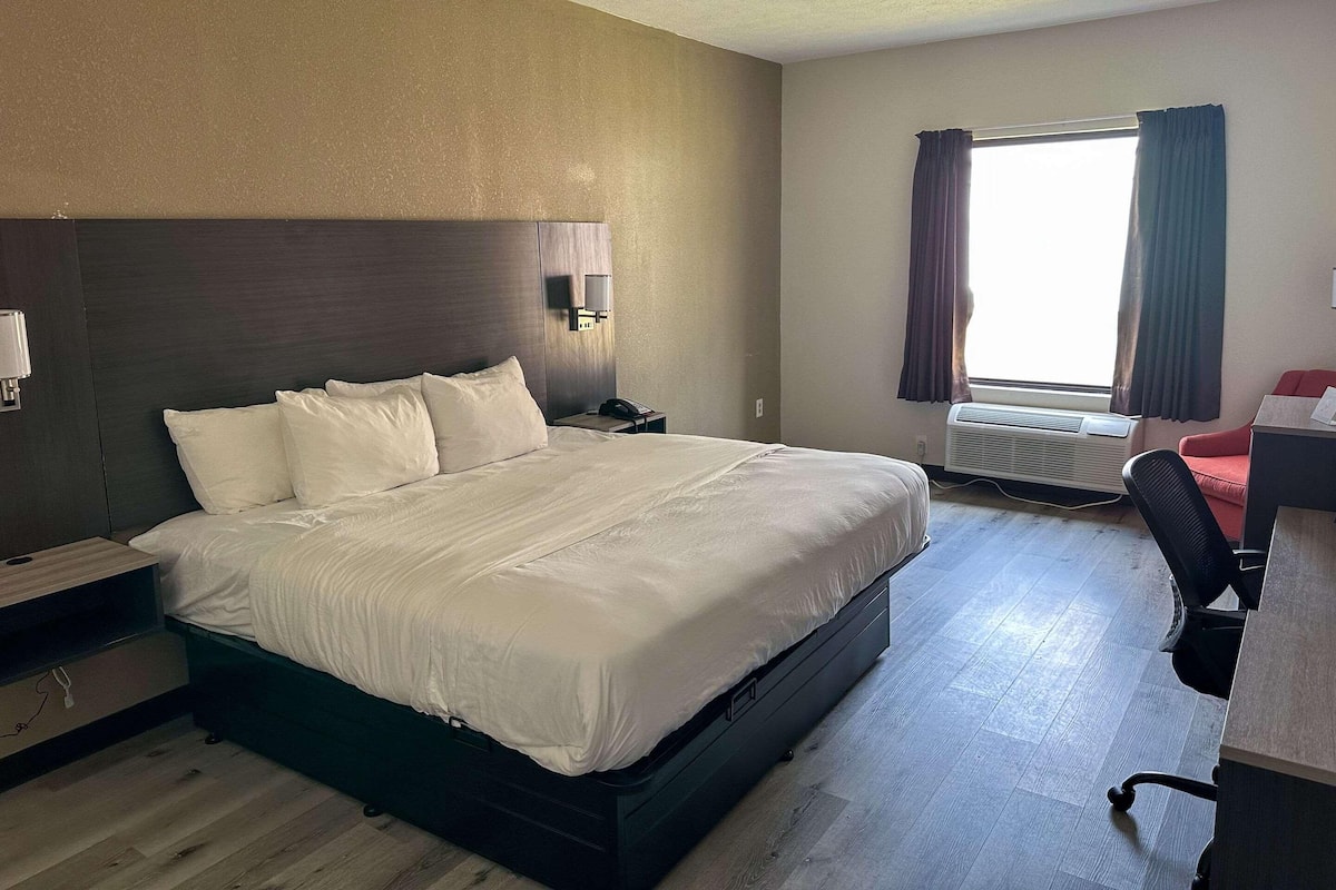 Une chambre d’hôtel comprenant un grand lit, un bureau, une chaise, une lampe de bureau, une table de chevet, une fenêtre avec des rideaux et un appareil de climatisation.