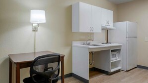 A compact kitchenette with a white countertop, a sink, a microwave, and a refrigerator. There is a chair and a table with a lamp.