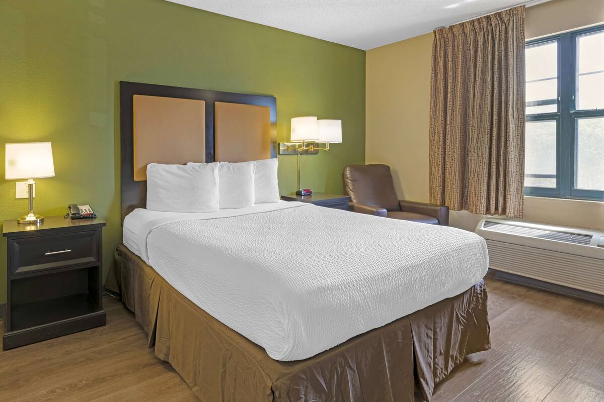 Habitación de hotel con una cama grande, una mesita de noche con lámpara, una silla y una ventana con cortinas.