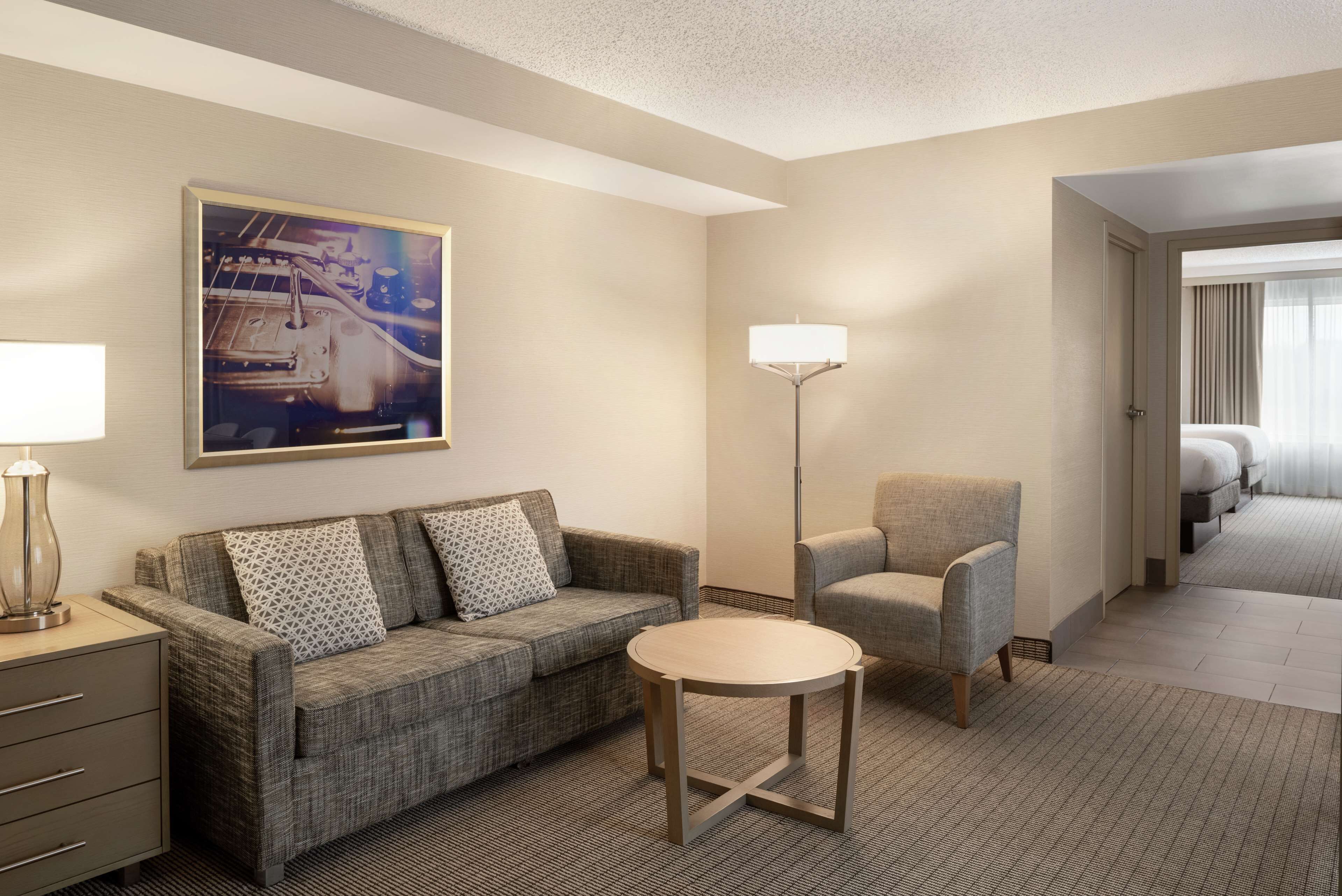 Habitación de hotel con un sofá gris, una mesita redonda pequeña, una lámpara de pie y un cuadro en la pared.