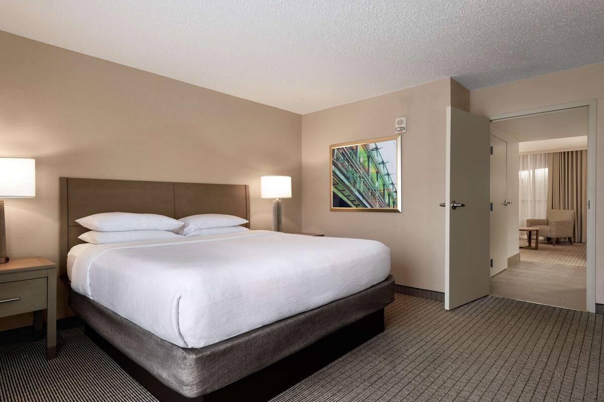 A hotel room with a large bed, a nightstand, a lamp, a painting on the wall, and a door leading to another room.