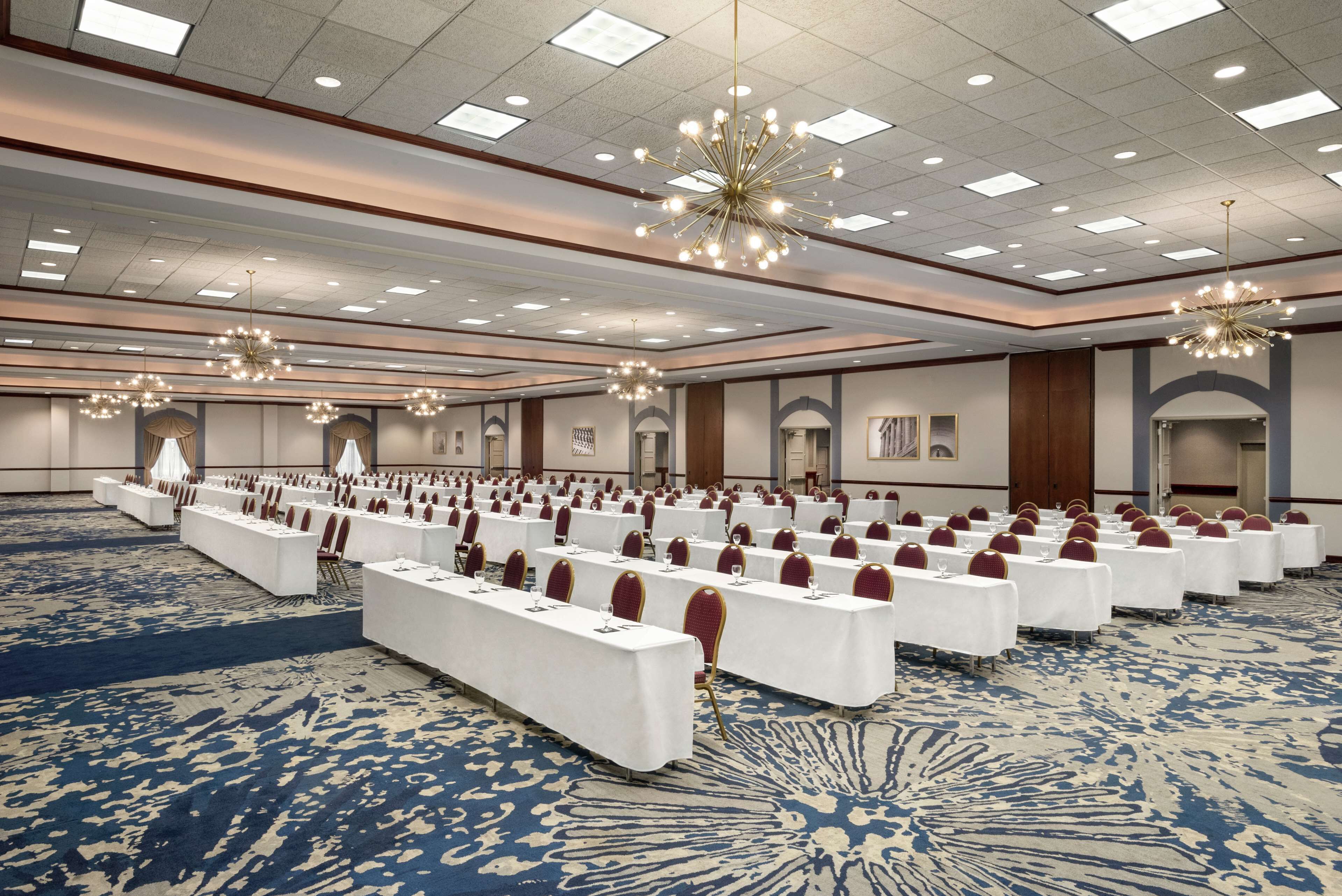 Un amplio salón de conferencias con filas de mesas y sillas, una alfombra estampada y modernos candelabros.