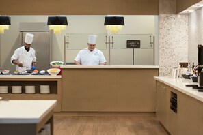 A modern kitchen with two chefs preparing food, a sign indicating the kitchen is ready for orders, and a clean, organized layout with wooden flooring and stainless steel appliances.
