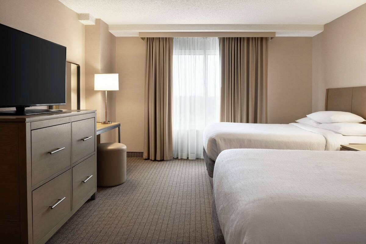 A hotel room with two beds, a television, a desk with a lamp, and a window with curtains.