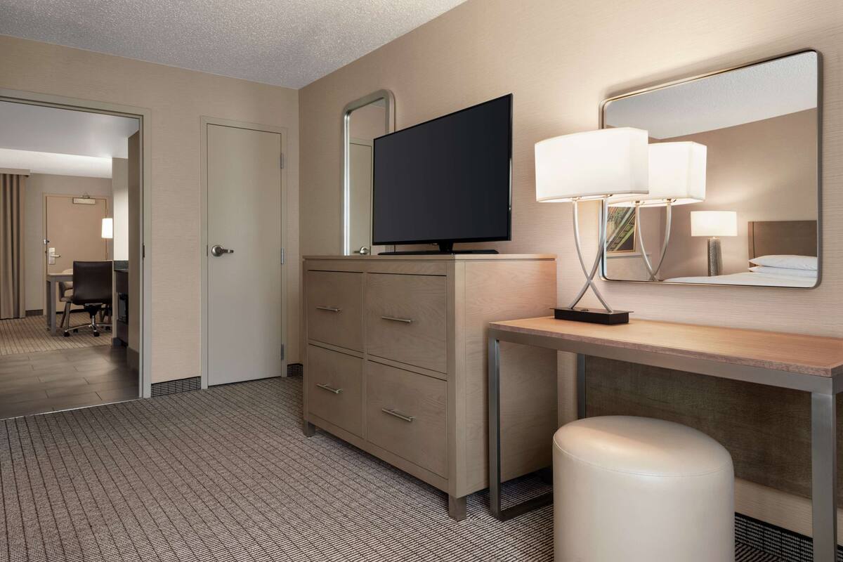 A hotel room with a flat-screen TV, a desk with a lamp, a mirror, and a chair.