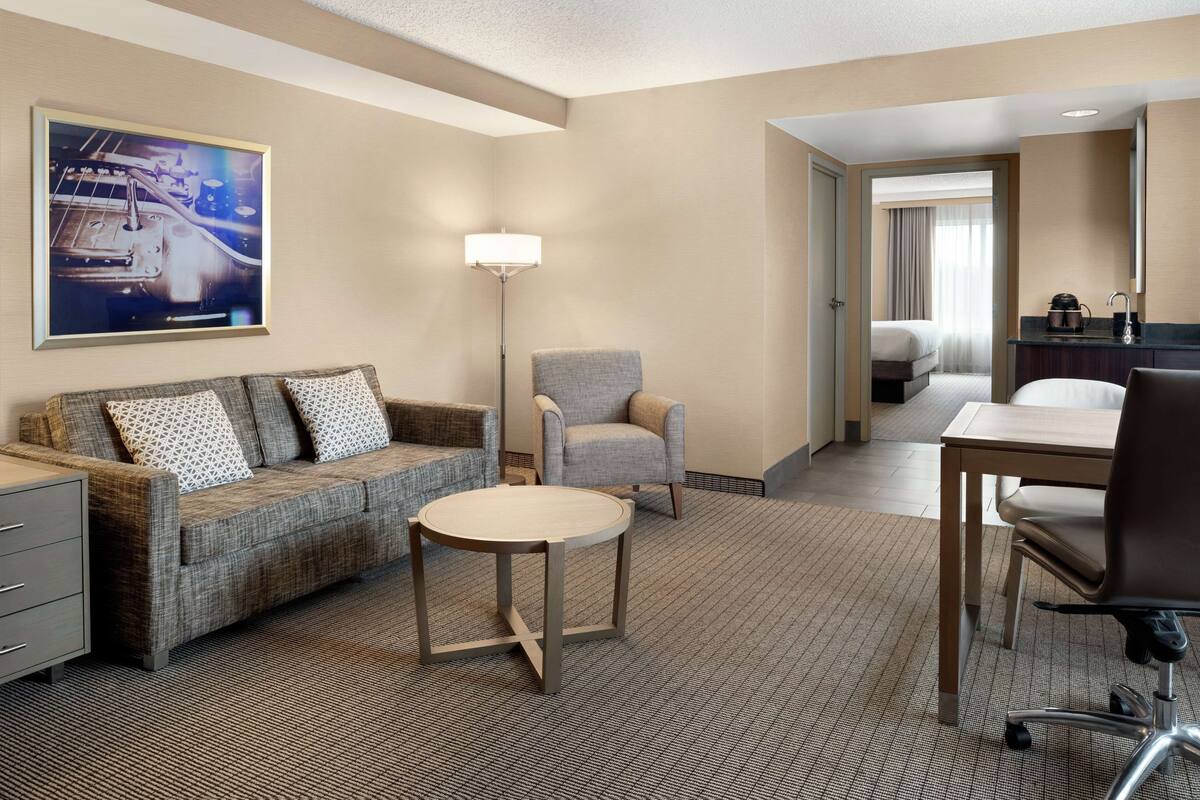 A hotel room with a sofa, armchair, small round table, and a desk with a chair. There is a painting on the wall and a bed in the background.