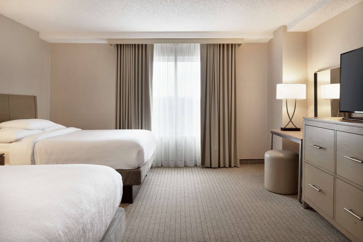 A hotel room with two beds, a desk with a lamp, a television, and a large window with curtains.