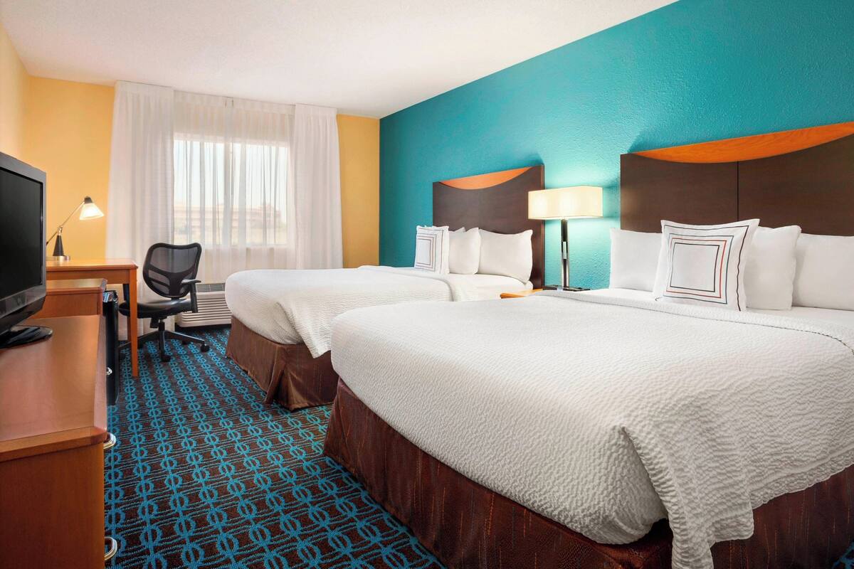 Habitación de hotel con dos camas, un escritorio con televisor, una silla y un ventanal con cortinas.