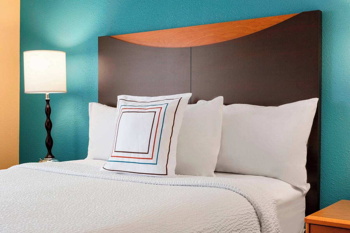 Una cama bien hecha con ropa blanca y una almohada decorativa de patrones geométricos, frente a una pared verde aguamarina y un cabecero de madera.