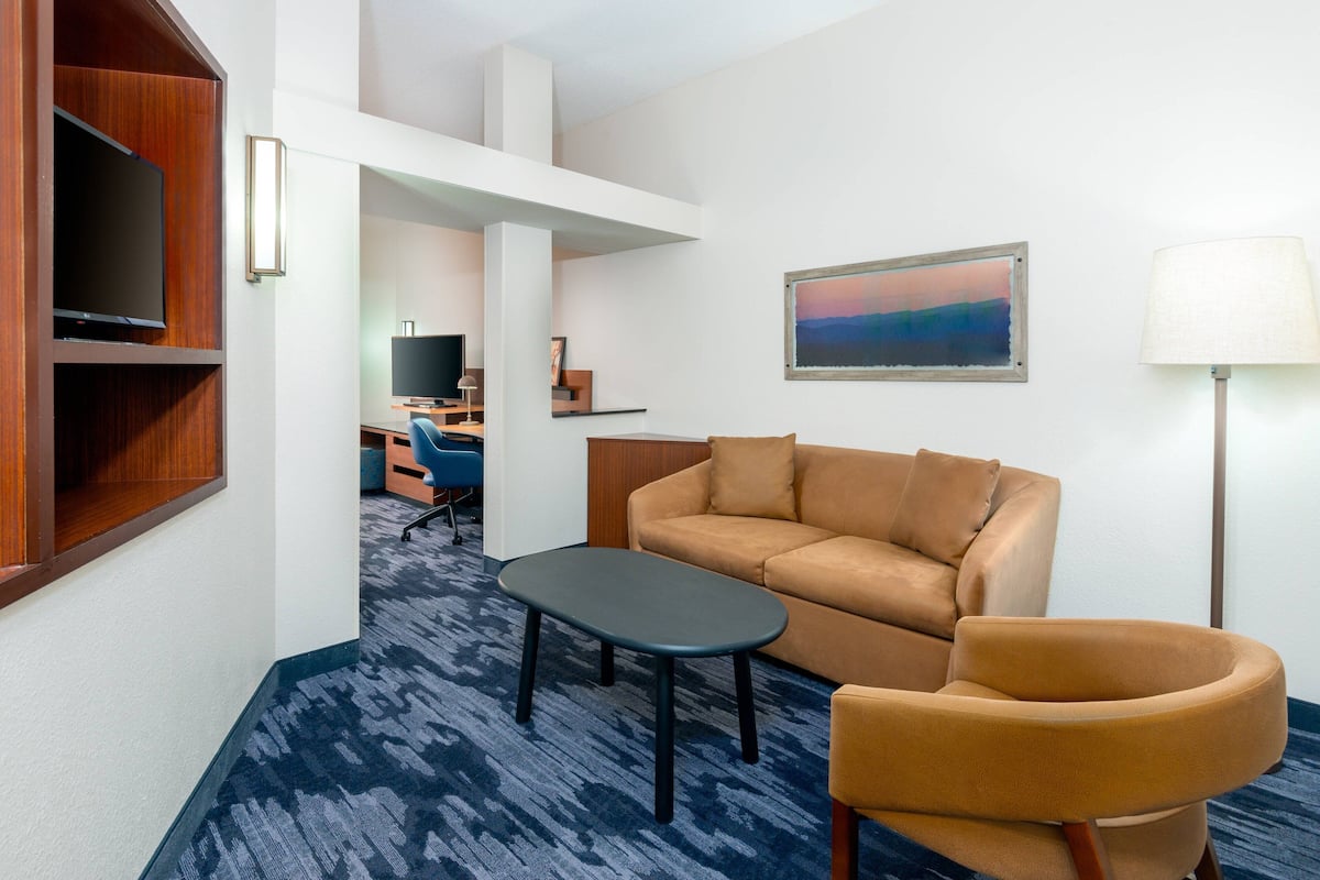 A modern hotel room with a sofa, armchair, and a small table. There is a TV mounted on the wall and a painting above the sofa.