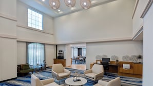 Un vestíbulo amplio con techo alto, una zona de estar central con sillones y una mesa de centro, un comedor al fondo y una recepción con computadora y objetos decorativos.