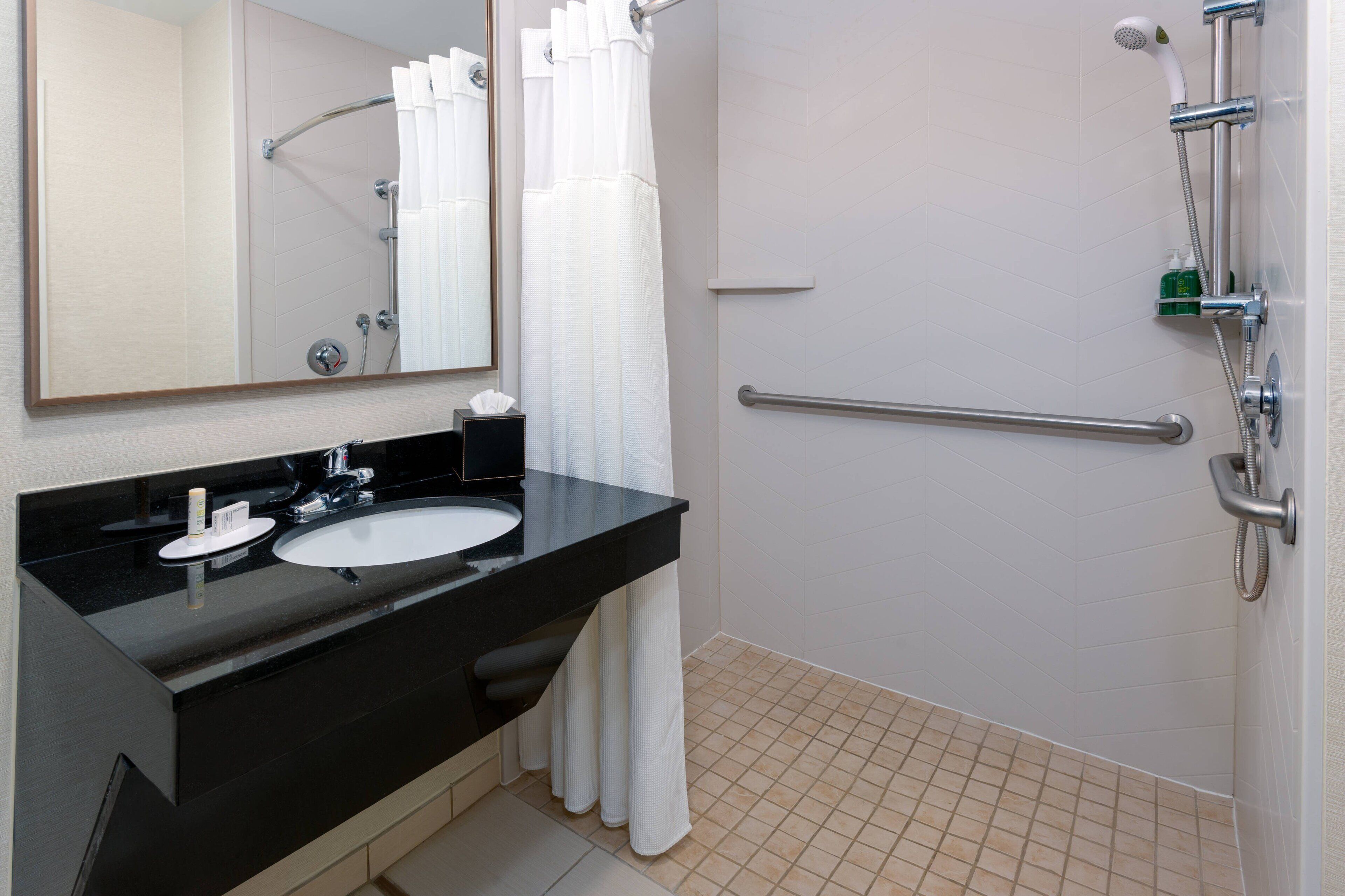 Un baño con un lavamanos negro, un espejo, ducha con pasamanos y suelo de baldosas blancas.