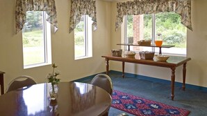 Un coin repas avec une table ronde, des chaises, un vase avec des fleurs, une table avec des paniers et une carafe, et une vue sur la verdure à travers les fenêtres.