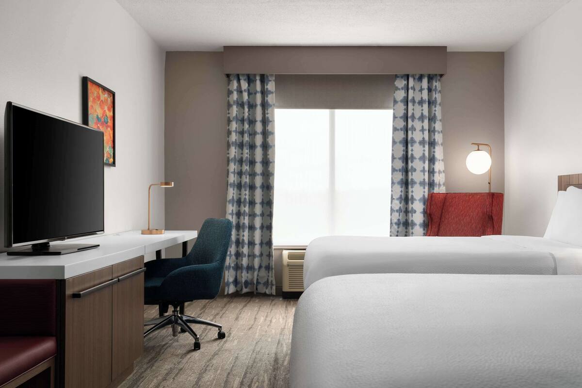 A hotel room with a bed, a desk with a TV, a chair, and a window with curtains.