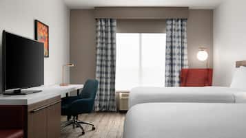 A hotel room with a flat-screen TV, a desk, a chair, a bed, a bedside table with a lamp, and a window with curtains.