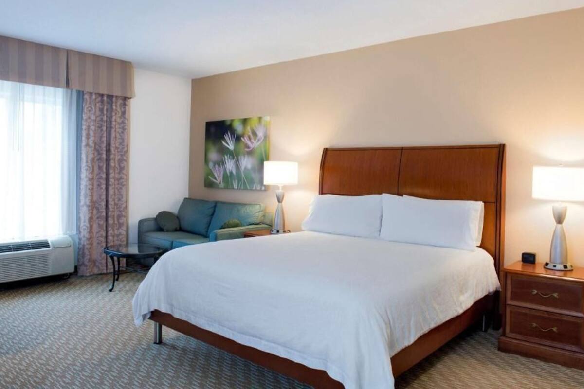 A hotel room with a large bed, a sofa, a nightstand with a lamp, and a painting on the wall.