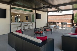 An outdoor seating area with wicker furniture and red cushions, a bar with stools, and a view of a brick wall. - Hilton Garden Inn Rock Hill (Rock Hill)