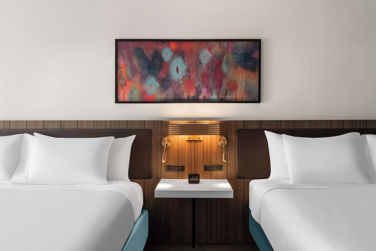 A hotel room with two beds, a wooden headboard, a small table with a clock, and a colorful abstract painting above the bed.