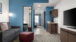 A modern hotel room with a grey sofa, red ottoman, and a TV on a wooden cabinet.