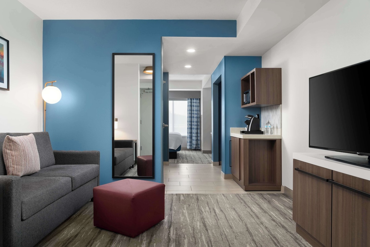 A modern hotel room with a grey sofa, red ottoman, and a TV on a wooden cabinet.