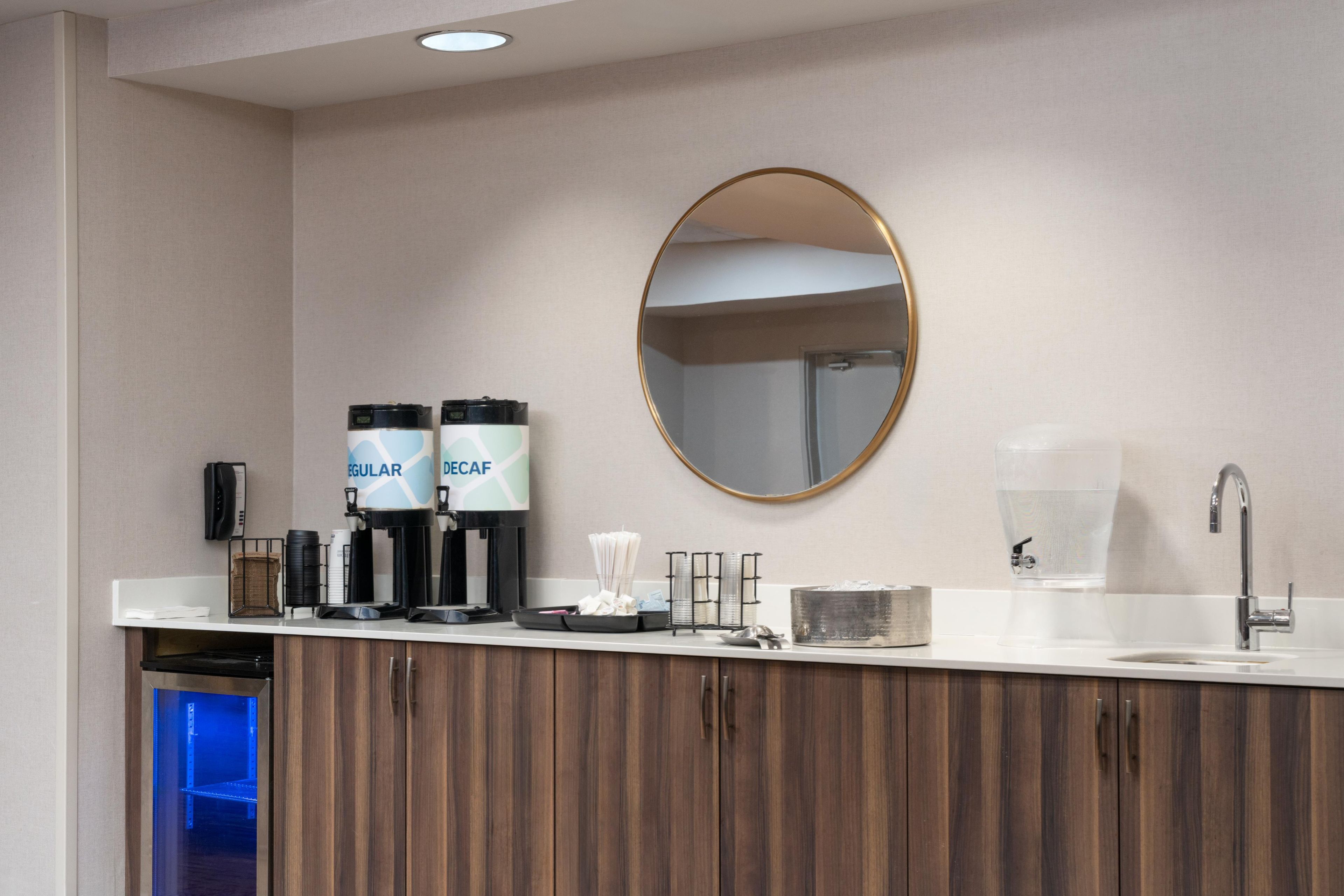 Casa de banho moderna com lavatório branco, espelho redondo e dois dispensadores de café identificados como 'Regular' e 'Descafeinado'.
