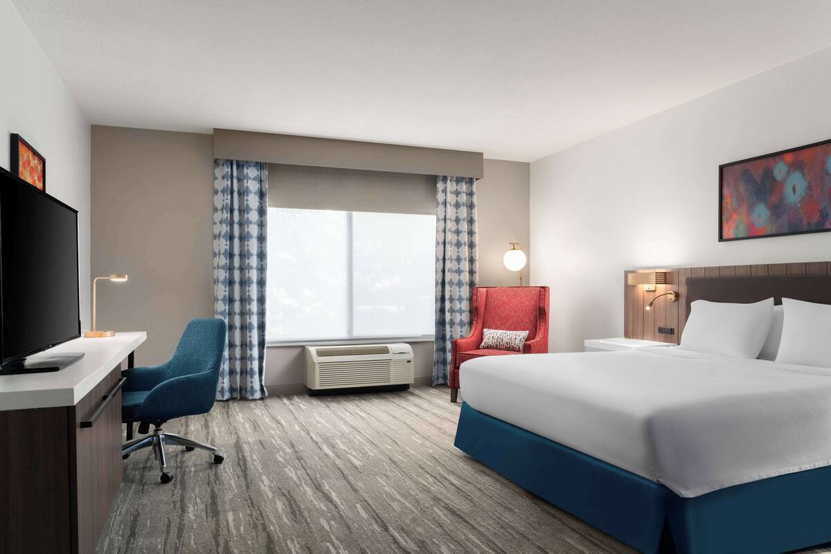 A hotel room with a bed, a desk with a computer, a chair, and a window with curtains.