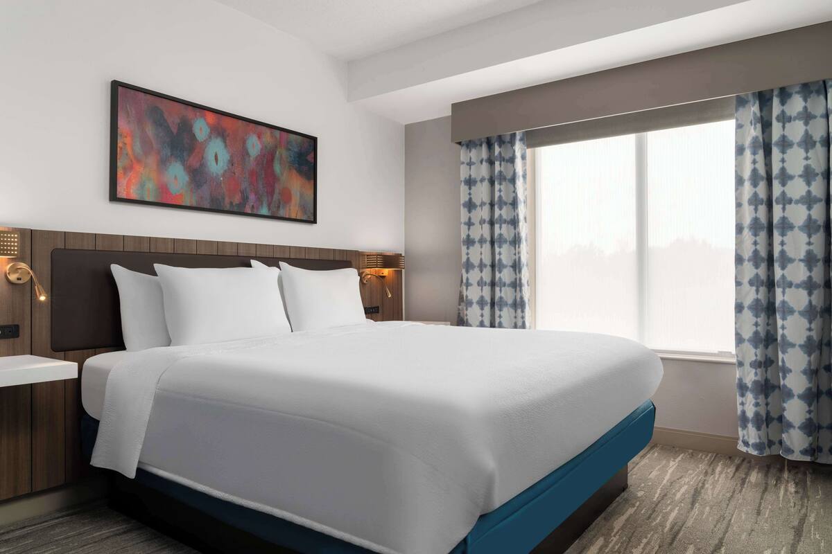 A hotel room with a large bed, a nightstand, a wall-mounted abstract painting, and a window with patterned curtains.