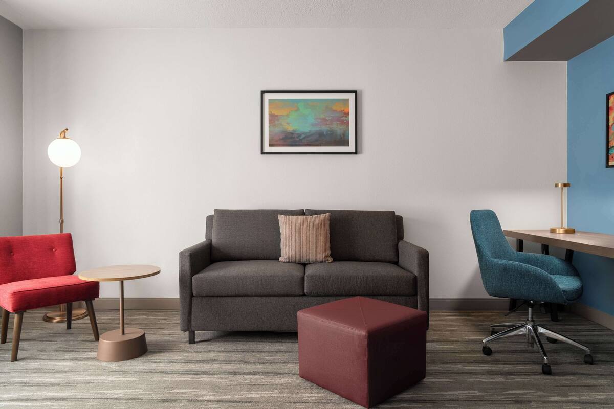 A modern living room with a grey sofa, a red armchair, a blue office chair, a small round table, and a red ottoman.