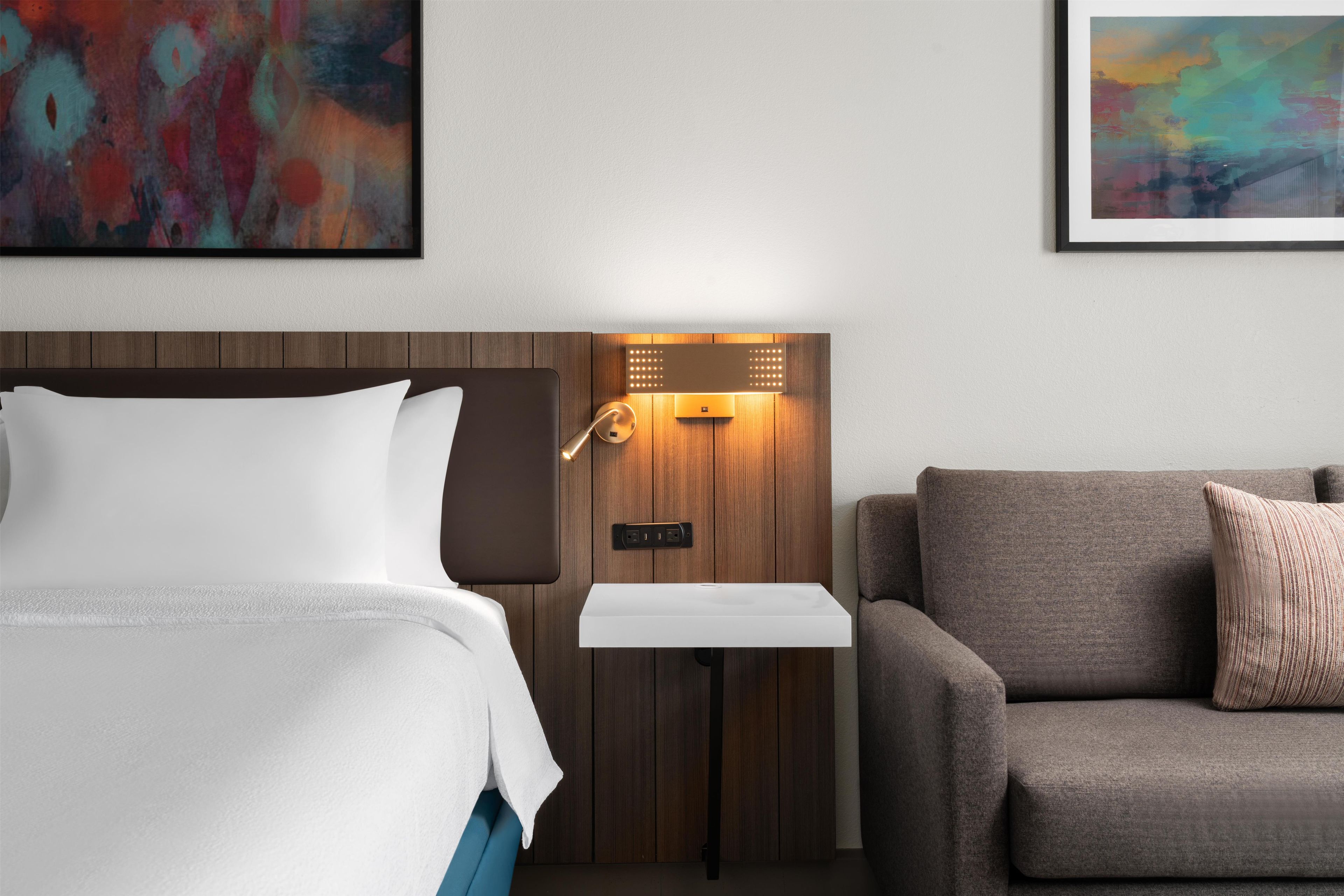 Una habitación de hotel con cama, sofá, mesita de noche y lámpara fijada a la pared.