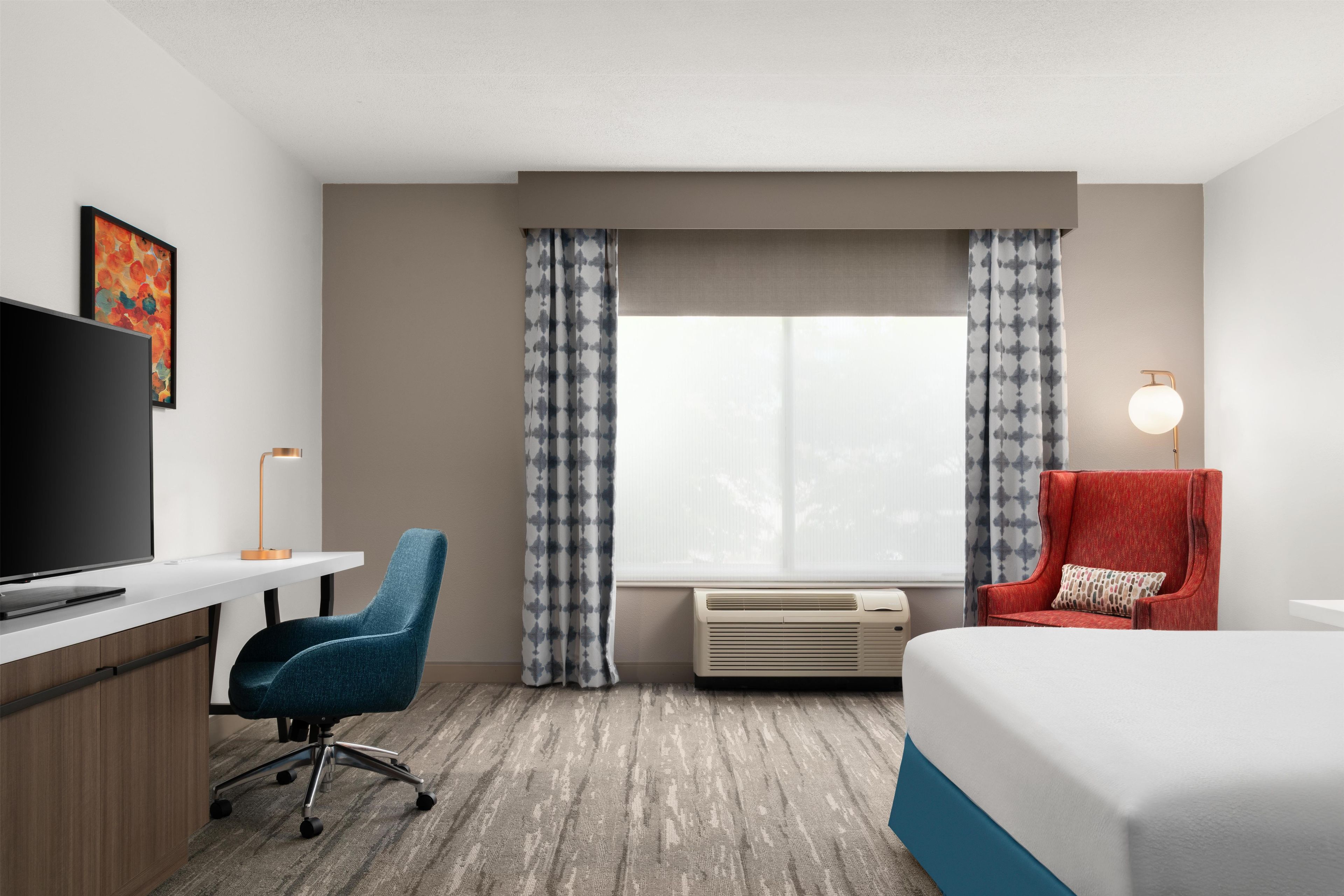 Een hotelkamer met een bed, bureau, stoel, televisie, airconditioning en een raam met gordijnen.