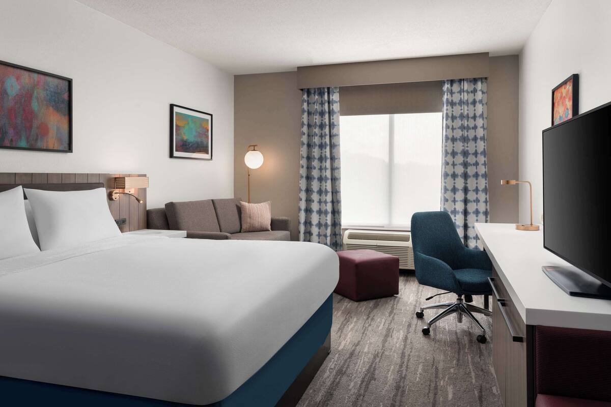 A hotel room with a large bed, a desk with a TV, a chair, a sofa, and a window with patterned curtains.